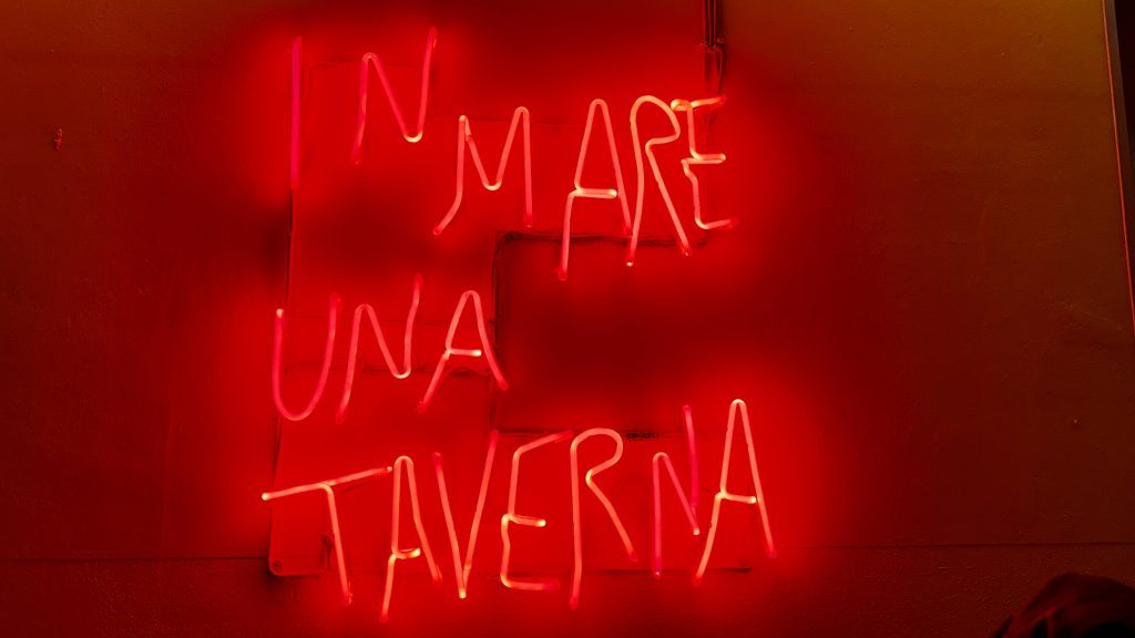 Neonlicht mit der Aufschrift "IN MARE UNA TAVERNA" auf einem roten Hintergrund.