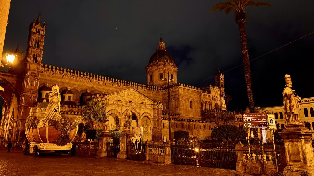 Nachtansicht der Kathedrale von Palermo mit kunstvoller Fassade, beleuchtet von Straßenlampen, und einer Statue in einem Boot im Vordergrund; umgeben von Palmen und Hinweisschildern.