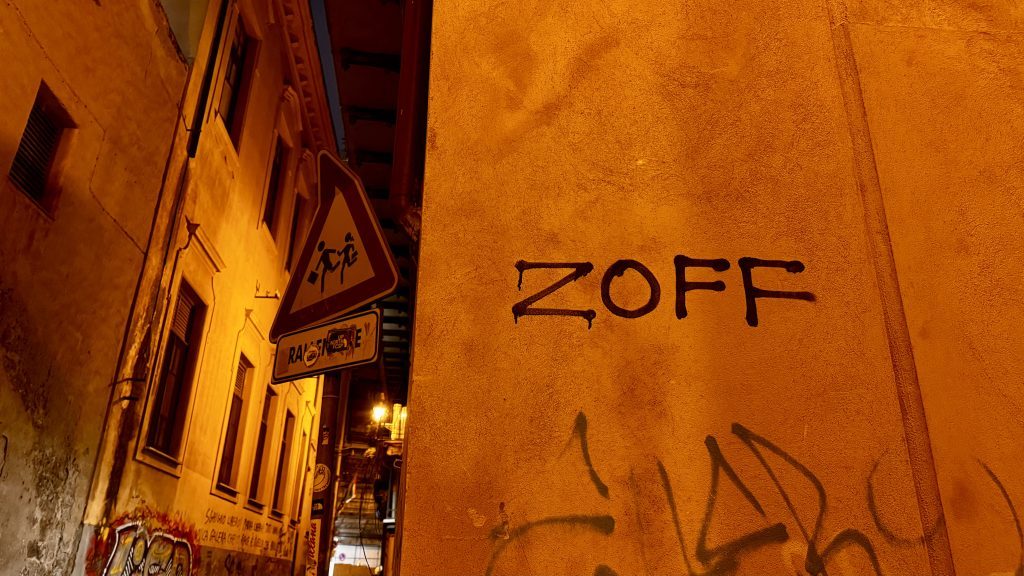 Das Bild zeigt eine schmale, beleuchtete Gasse mit orangefarbenen Wänden, an der eine Wand mit dem Schriftzug "ZOFF" zu sehen ist. Im Hintergrund steht ein Verkehrsschild, das auf laufende Personen hinweist.