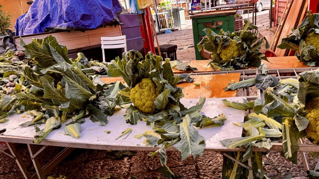 Ein Tisch auf einem Markt ist mit frischem Blumenkohl und Gemüse umgeben, während Blätter und Stängel von dem Gemüse herumliegen. Im Hintergrund sind Strukturen und Möbel eines Marktes zu erkennen.