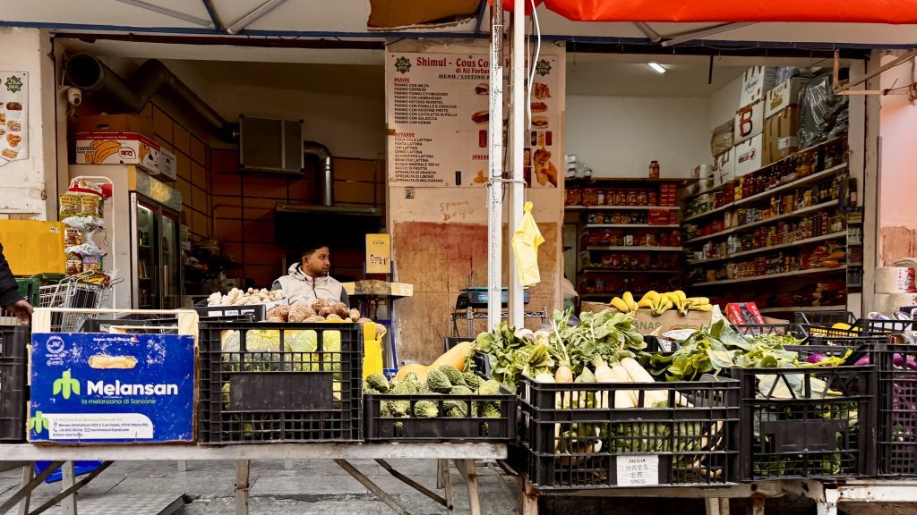 Ein Marktstand mit verschiedenen Obst- und Gemüsearten wie Brokkoli, Kürbis und Bananen ist im Vordergrund zu sehen. Im Hintergrund steht ein Mann in einem Kiosk, der Snacks und Getränke verkauft.
