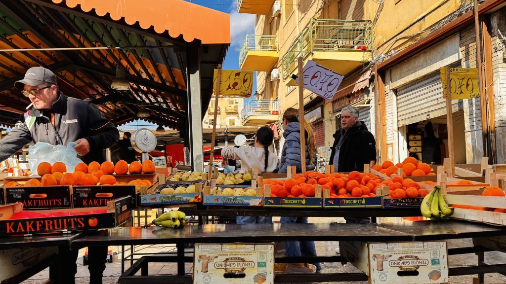 Ein Marktstand mit orangefarbenen Früchten und Bananen, an dem ein Verkäufer mit einer Blauen Tüte arbeitet. Im Hintergrund sind Kunden zu sehen, sowie Stellschilder mit Preisen für Obst.