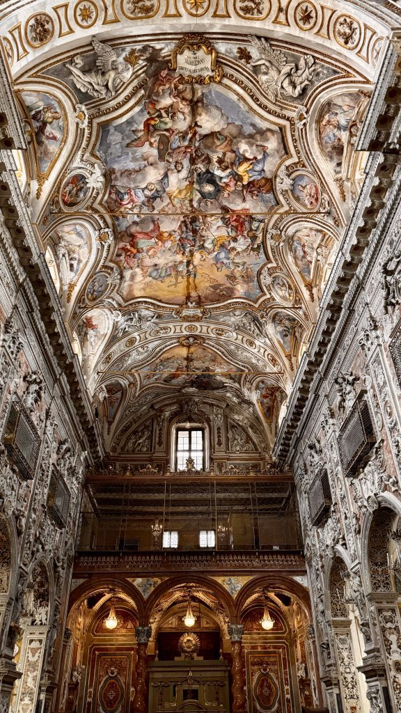Blick auf die kunstvoll gestaltete Decke und die reich verzierten Wände eines historischen Innenraums, vermutlich einer Kapelle oder Kirche, mit eindrucksvollen Fresken und goldenen Akzenten sowie einem Fenster in der oberen Etage.