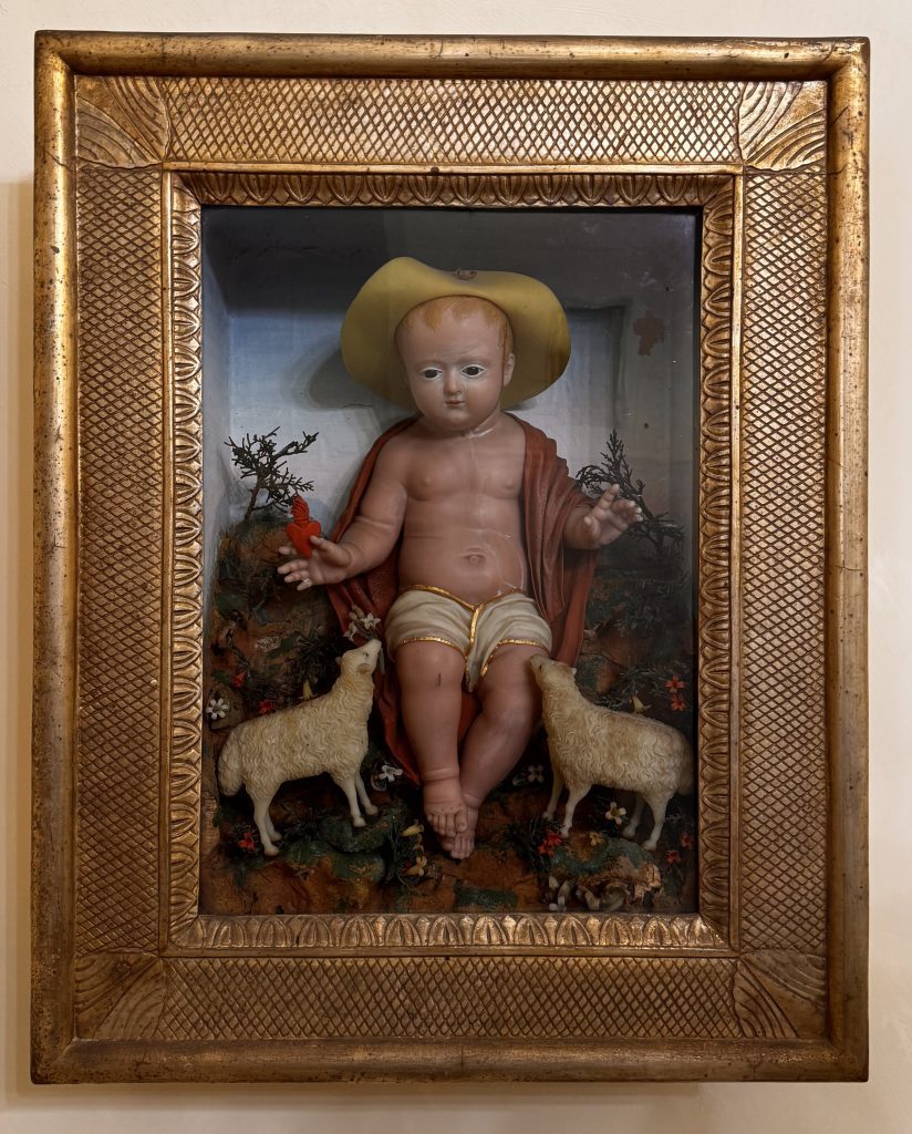 Eine religiöse Figur eines nackten, sitzenden Kindes mit einem gelben Hut, umgeben von zwei Schafen und bunten Blumen in einem goldverzierten Rahmen. Der Hintergrund zeigt eine ländliche Szenerie.