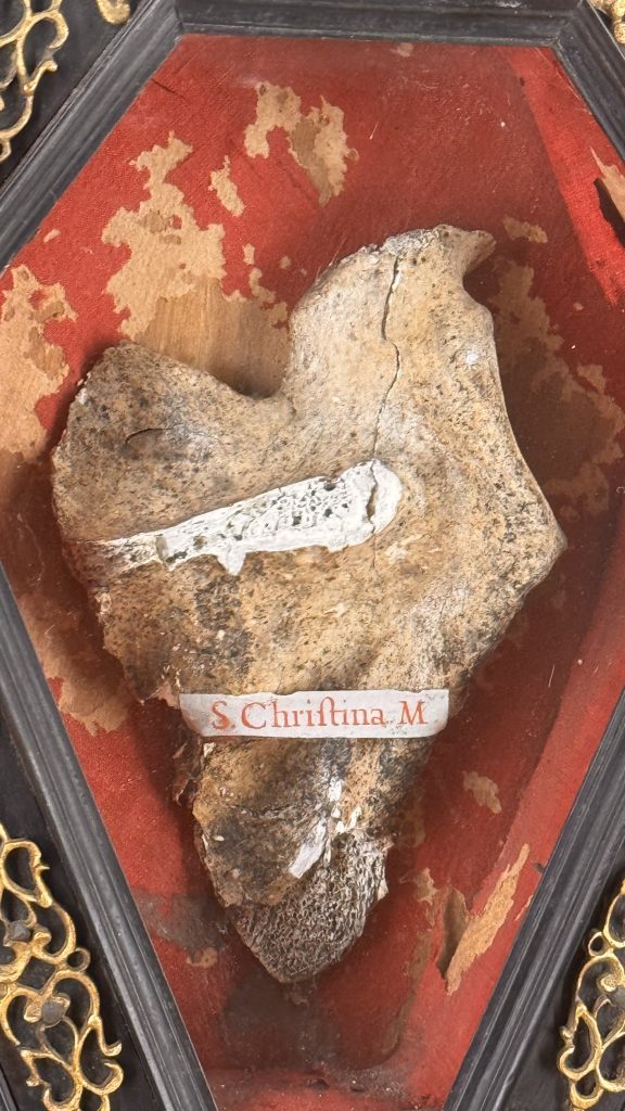 Eine herzförmige Reliquie aus alter Kleidung oder Holz ist in einem runden Rahmen auf rotem Hintergrund präsentiert. Darunter steht in einer weißen Schrift "S. Christina M".
