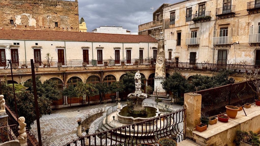 Blick auf einen Innenhof mit einem Brunnen und einer Statue in der Mitte, umgeben von grünen Bäumen und alten Gebäuden mit Fensterläden. Die Fassade der Gebäude ist teils abblätternd, und der Himmel ist bewölkt.