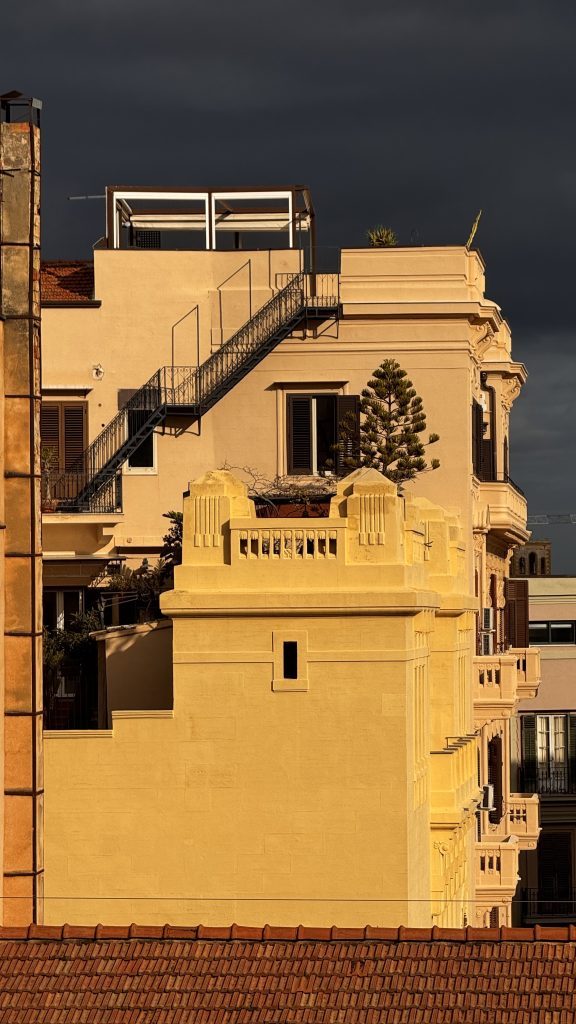 Das Bild zeigt einen Teil eines Gebäudes mit einer gelben Fassade und einem dekorativen Balkon, flankiert von einem weiteren Gebäude mit einer Stahlfeuertreppe. Der Himmel ist düster und bewölkt, während die Sonne einen Teil der Fassade beleuchtet.