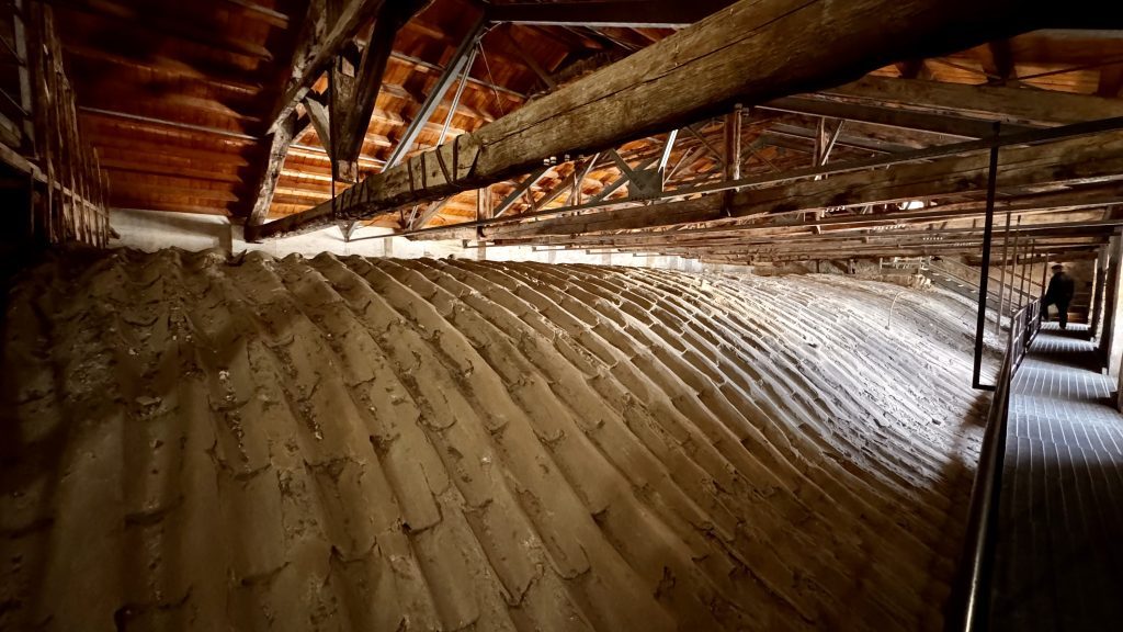 Blick auf eine gewölbte, bepflasterte Fläche in einem Dachraum mit Holzträgern und einem Gehweg, der an der rechten Seite verläuft; im Hintergrund ist eine Person zu sehen.
