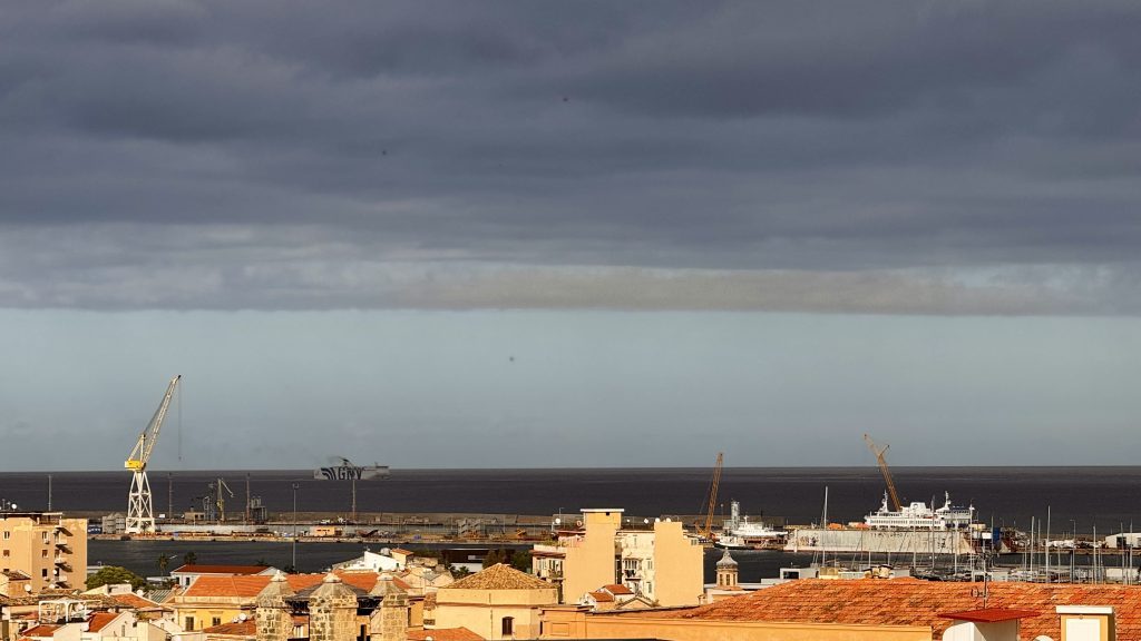 Blick auf einen Hafen mit Schiffen und Kränen, umgeben von Häusern mit roten Dächern unter einem bewölkten Himmel. Im Hintergrund sind verschiedene Bauarbeiten sichtbar.