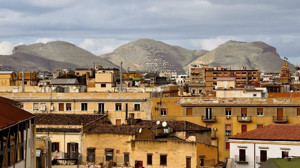 Stadtansicht mit flachen, grauen Hügeln im Hintergrund und mehrstöckigen, überwiegend gelben und beige Häusern im Vordergrund. Auf den Dächern sind Satellitenschüsseln zu sehen, und der Himmel ist bewölkt.