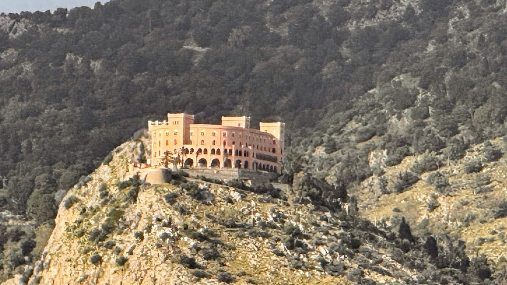 Eine historische, terrakottafarbenen Villa steht auf einem Hügel in einer bergigen Landschaft, umgeben von Bäumen und Felsen. Im Hintergrund sind die sanften Konturen der Berge sichtbar.