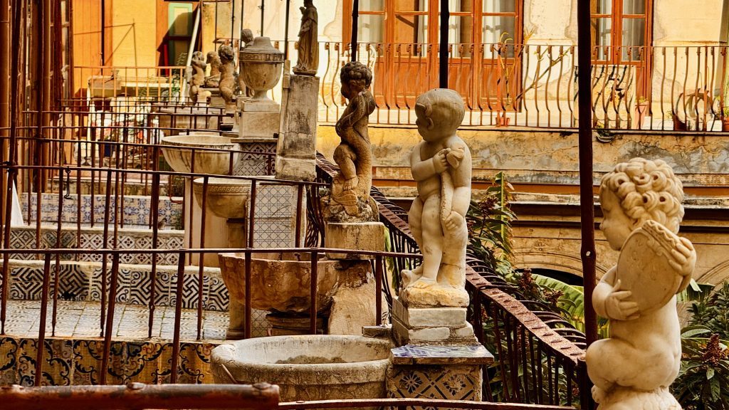 Ein Innenhof mit mehreren Marmorstatuen von Kindern und Engeln, umgeben von rustikalen Metallgeländern und dekorativen Fliesen. Im Hintergrund sind vergitterte Fenster und Pflanzen zu sehen.
