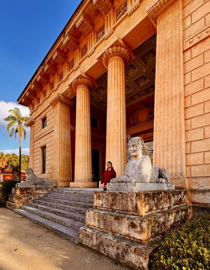 Ein Gebäude im antiken Stil mit Säulen steht im Vordergrund, umgeben von Stufen. Eine Person in rotem Oberteil steht neben einer steinernen Sphinx und einer liegenden Löwenstatue.