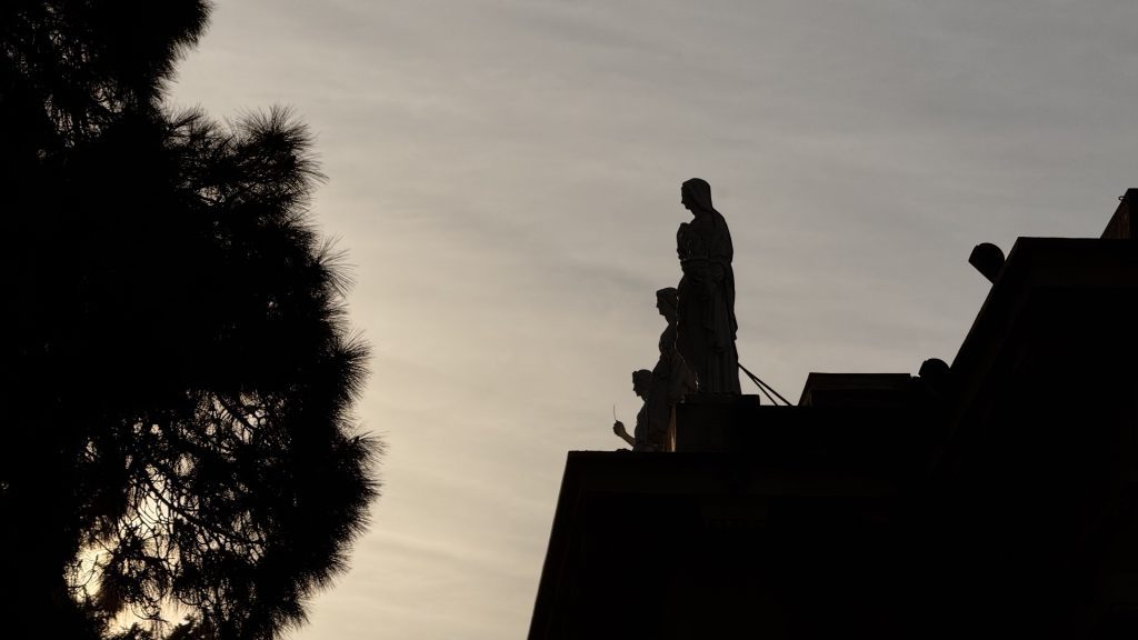 Silhouette von drei Statuen auf einem Dach, während ein Baum im Vordergrund sichtbar ist. Der Himmel hat eine sanfte, graue Färbung.