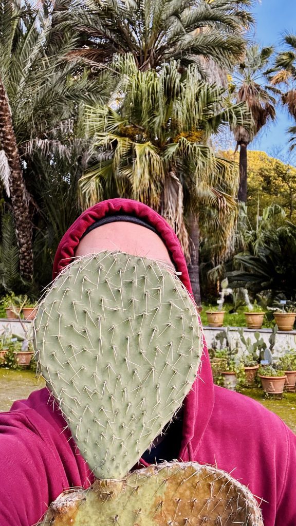 Eine Person mit Kapuzenpulli hält ein großes Kaktusblatt vor ihr Gesicht, während sie in einem Garten mit vielen Palmen und Töpfen voller Pflanzen steht.