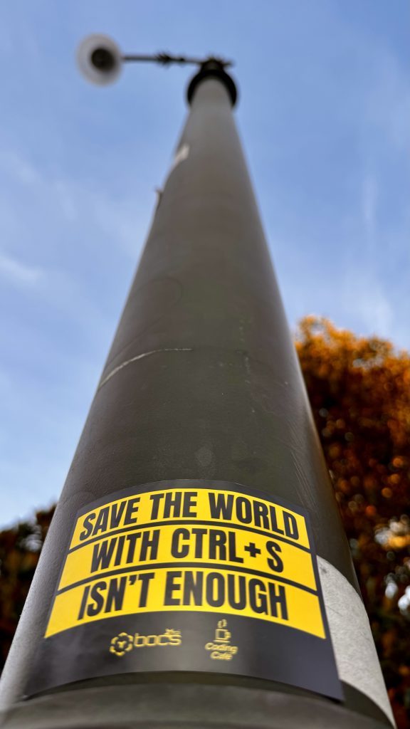 Ein Straßenlaternenmast mit einem auffälligen gelben Aufkleber, der den Text "SAVE THE WORLD WITH CTRL+S ISN'T ENOUGH" zeigt. Der Hintergrund zeigt einen klaren Himmel und teilweise grün gefärbte Bäume.