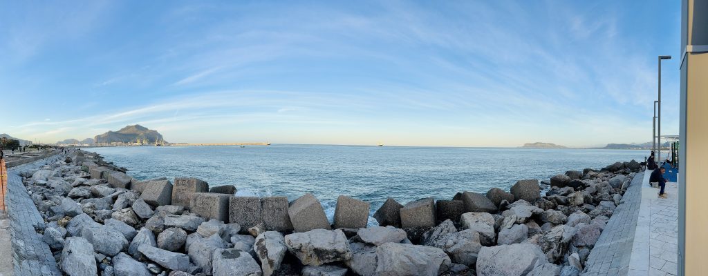 Panoramablick auf das Meer mit einer felsigen Küste im Vordergrund. Im Hintergrund sind Gebirgen und der Hafenvisible, während einige Menschen auf Bänken sitzen und spazieren.