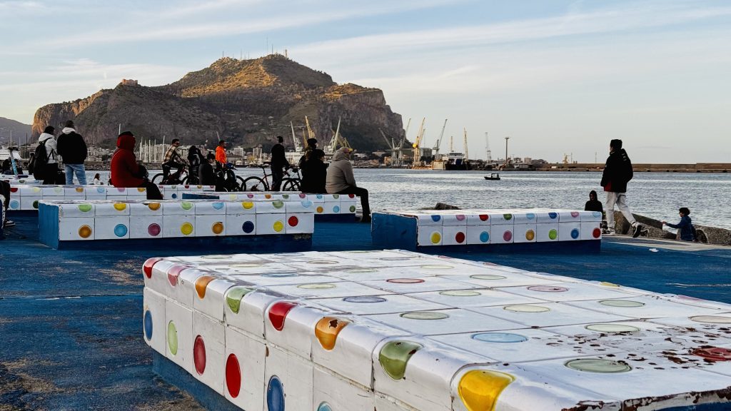 Menschen sitzen und stehen auf bunten Sitzgelegenheiten am Hafen, während im Hintergrund kranartige Gebäude sichtbar sind und Boote auf dem Wasser fahren. Der Blick reicht über das Meer auf einen bewaldeten Hügel mit einer Festung.