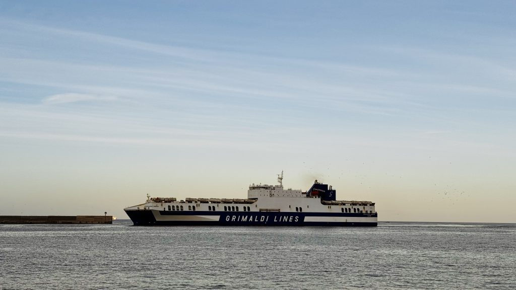 Ein großes Fährschiff der Grimaldi Lines fährt auf dem ruhigen Wasser, im Hintergrund sind ein blauer Himmel und eine Küstenlinie mit einem kleinen Hafen zu sehen.