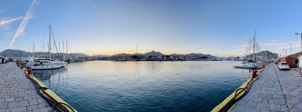 Blick auf einen Hafen mit mehreren Segelbooten, die am Steg festgemacht sind, während die Sonne hinter den Bergen aufgeht und den Himmel in sanften Farben erhellt. Der Steg ist mit roten Pollern sowie Schläuchen ausgestattet, und im Hintergrund sind Gebäude und Berge sichtbar.