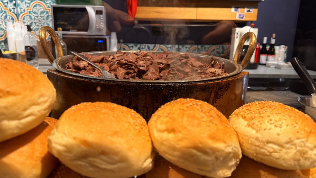 Ein großer Topf mit dampfendem, geschnittenem Fleisch steht in der Mitte des Bildes, während im Vordergrund mehrere frische, sesam-bestreute Brötchen angeordnet sind. Im Hintergrund sind Küchengeräte und Flaschen sichtbar.