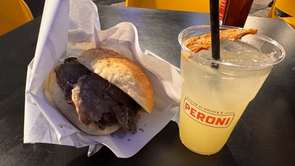 Ein belegtes Brötchen mit dünn geschnittenem Fleisch liegt in einem Papier, daneben steht ein Becher mit einem klaren Getränk, garniert mit einer Trockenfruchtscheibe. Im Hintergrund sind gelbe Stühle und ein Tisch zu sehen.