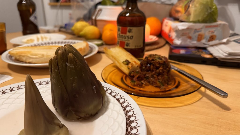Ein Tisch ist gedeckt mit verschiedenen Speisen, darunter zwei Artischockenhälften, ein Teller mit Fladenbrot, ein glasierter Teller mit einer Mischung aus Gemüse und Fleisch, eine Flasche Getränk sowie Obst im Hintergrund.