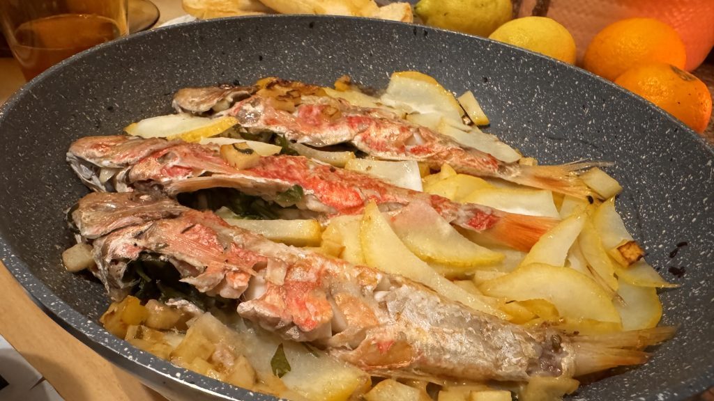 Ein flacher Topf mit drei gebratenen Fischen, umgeben von dünn geschnittenem Gemüse, darunter Zwiebeln und Kartoffeln. Im Hintergrund sind verschiedene Obstsorten wie Äpfel und Orangen sichtbar.