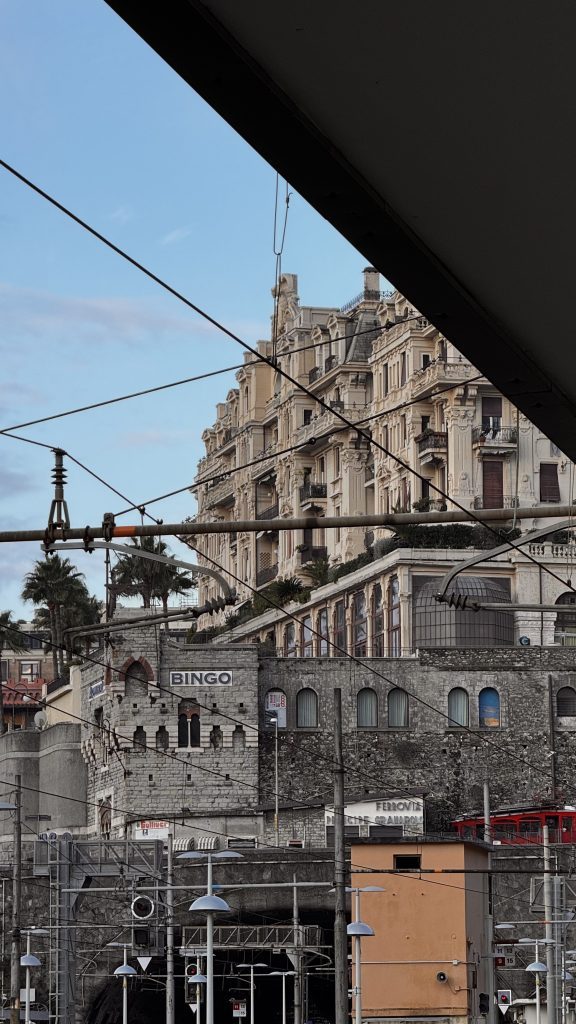 Blick auf ein historisches Gebäude mit einem eleganten Fassadendesign, das entlang eines Hanges steht, umgeben von modernen Infrastrukturen und Oberleitungen. Im Vordergrund sind Teile eines Bahnhofs mit verschiedenen Schildern und Straßenbeleuchtung sichtbar.