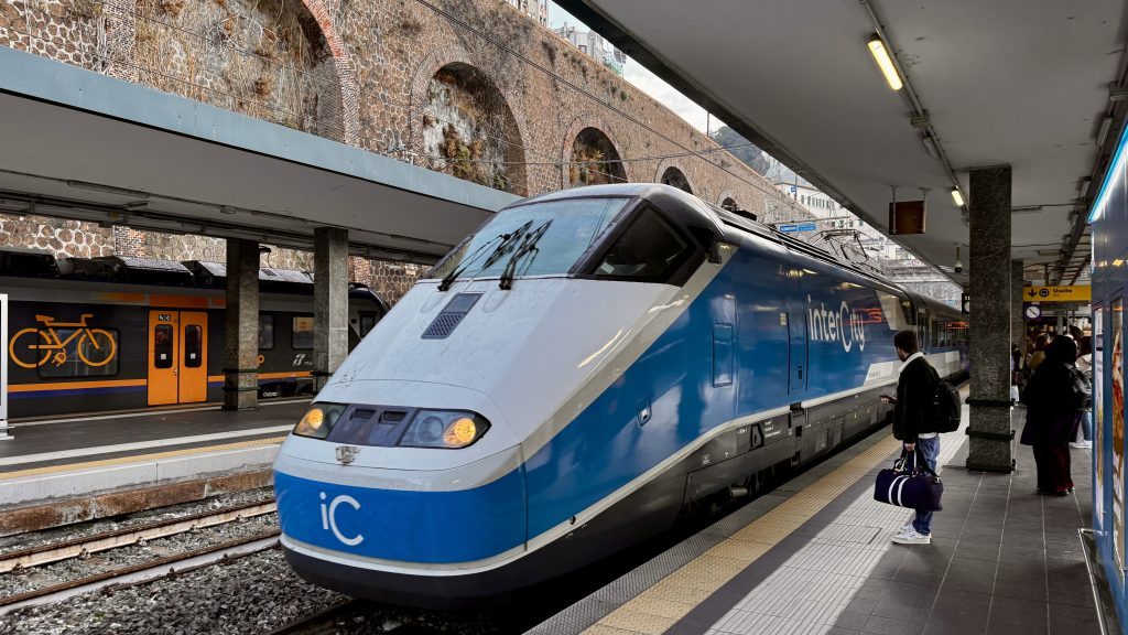 Zug der Intercity-Linie steht an einem Bahnhof, während Fahrgäste auf dem Bahnsteig warten. Im Hintergrund ist ein weiterer Zug sichtbar, dessen Türen geöffnet sind.