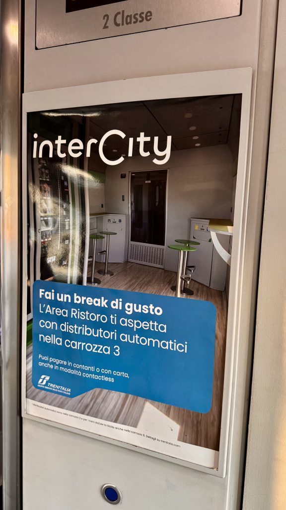 Ein Werbeplakat für den InterCity-Zug zeigt eine großzügige Snack- und Getränke-Ecke mit Automaten in einem modernen Zugabteil. Der Text lädt die Passagiere ein, sich im Bereich der Wagen 3 zu bedienen und informiert über Zahlungsmöglichkeiten.
