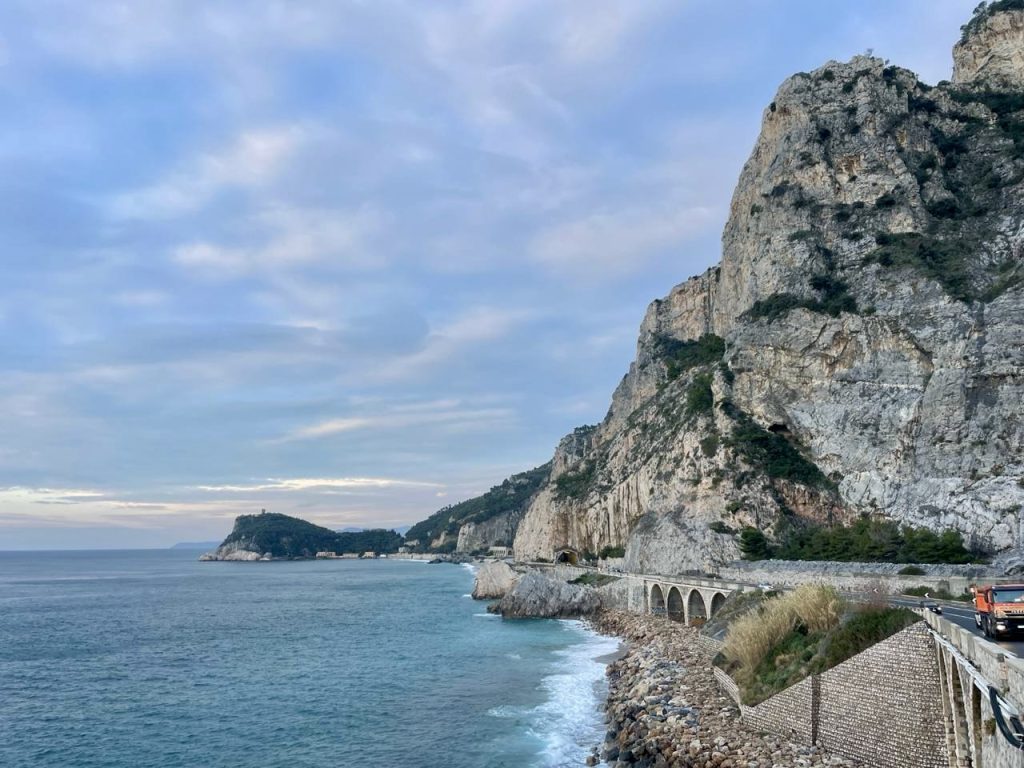 Blick auf eine Küstenstraße, die sich entlang eines felsigen Berges erstreckt, mit klarem Wasser und sanften Wellen im Vordergrund. Am Horizont sind bewaldete Hügel und ein kleiner Hügel mit einer Ruine sichtbar, während ein orangefarbener Lkw auf der Straße fährt.
