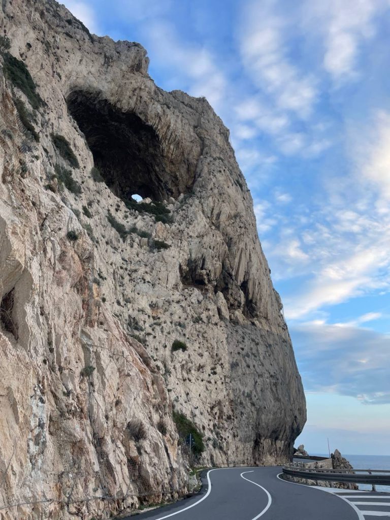 Blick auf eine kurvenreiche Straße, die entlang einer steilen Felswand verläuft, die ein großes rundes Loch aufweist. Der Himmel ist blau mit einigen Wolken.