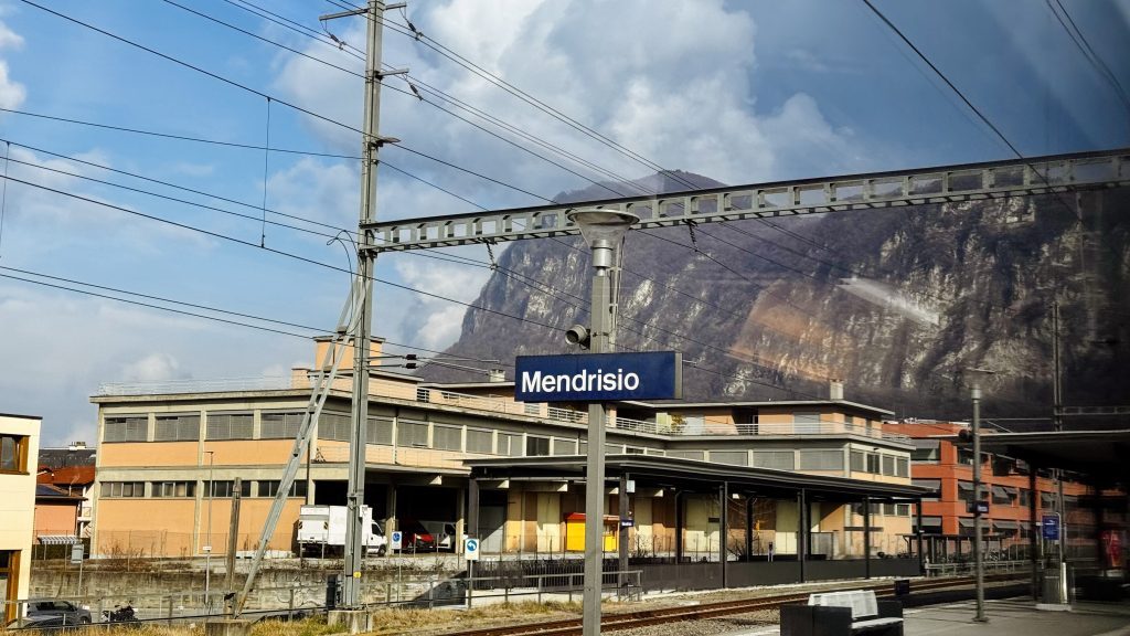 Bahnhof Mendrisio mit einem modernen Gebäude im Vordergrund, umgeben von Gleisen und elektrischen Leitungen, im Hintergrund ist ein bewaldeter Hügel zu sehen. Der Himmel ist teilweise bewölkt.