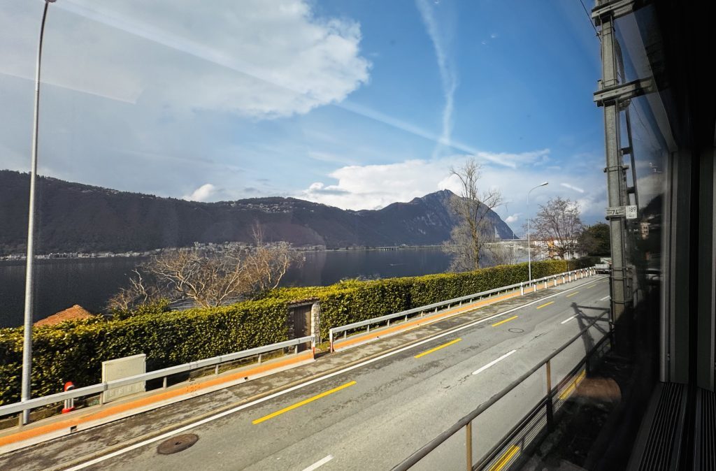 Eine Aussicht auf einen klaren See, umgeben von Bergen und bewaldeten Hängen, mit einer Straße im Vordergrund, auf der sich ein Fahrzeug befindet. Am Ufer sind Bäume und eine Hecke sichtbar, während Wolken am Himmel verteilt sind.