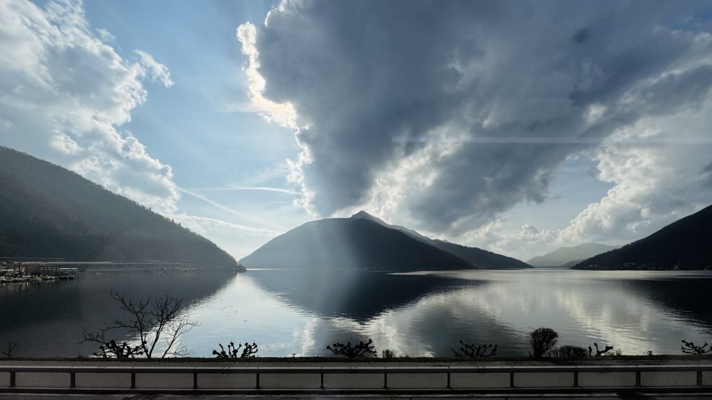 Blick auf einen ruhigen See, umgeben von Bergen und bewölktem Himmel, mit reflektierenden Wasseroberflächen und vereinzelten Bäumen am Ufer. Die Sonne bricht durch die Wolken und erzeugt Lichtstrahlen, die auf das Wasser fallen.