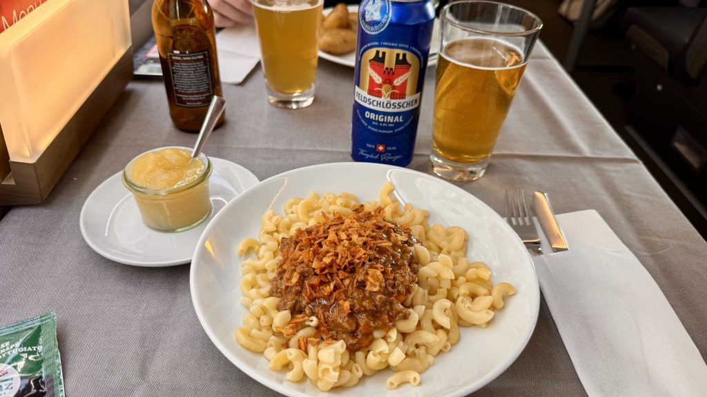 Ein Teller mit gekochten Hörnchen-Nudeln, garniert mit einer herzhaften Fleischsauce und Röstzwiebeln, steht neben einem Glas Apfelmus auf einem Tisch. Im Hintergrund sind mehrere Biergläser und eine Bierdose zu sehen.