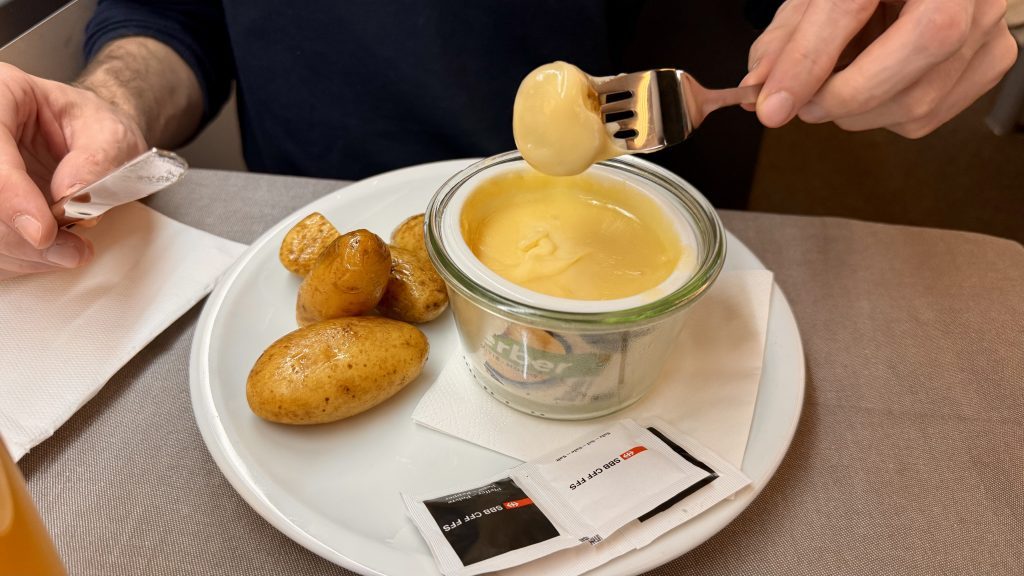 Eine Person hält eine Gabel mit einer cremigen Speise, die aus einem Glasbehälter entnommen wird, während auf einem Teller kleine, gekochte Kartoffeln liegen. Im Hintergrund sind zwei Päckchen mit Gewürzen sichtbar.