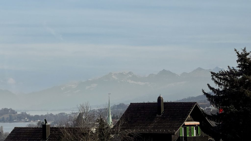 Blick auf eine hügelige Landschaft mit Häusern im Vordergrund und schneebedeckten Bergen im Hintergrund, der Himmel ist leicht bewölkt und hat einen bläulichen Farbton. Ein Baum und ein Kirchturm sind ebenfalls sichtbar.