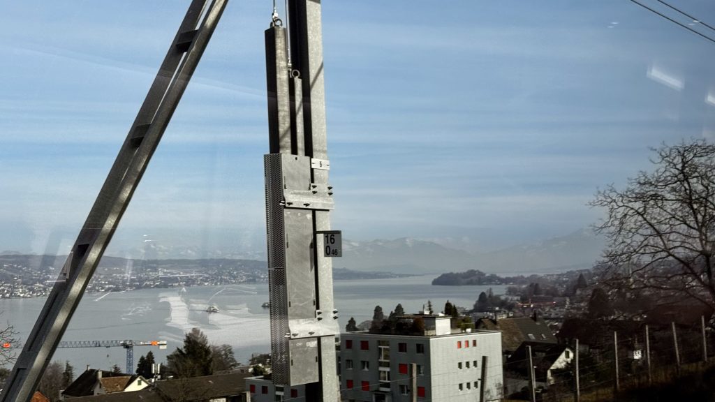 Ein Blick auf einen See mit Bergen im Hintergrund, während ein stehendes, metallisches Baugerüst im Vordergrund sichtbar ist. In der Nähe stehen moderne Wohngebäude und ein Kran ist ebenfalls zu sehen.