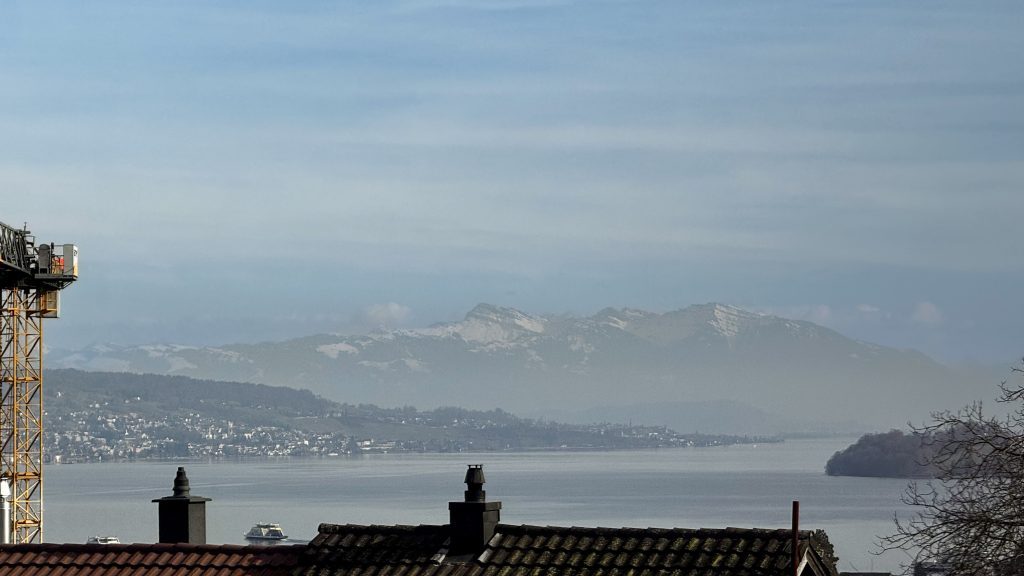 Blick auf einen ruhigen See mit Bergen im Hintergrund. Im Vordergrund sind Teile von Dächern und Schornsteinen zu sehen.