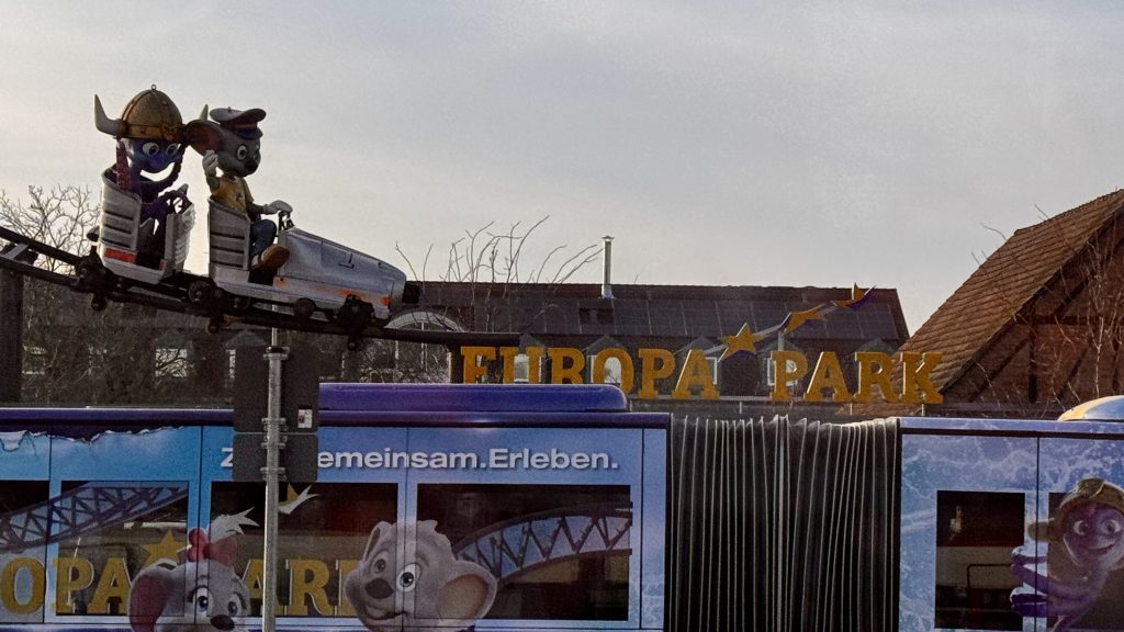 Zwei animierte Figuren in einem kleinen Zug auf einer Schiene, einer trägt einen Helm und der andere einen Hut, im Hintergrund steht das Logo von Europa Park. Ein lila Bus mit Illustrationen von cartoonhaften Tieren fährt darunter.