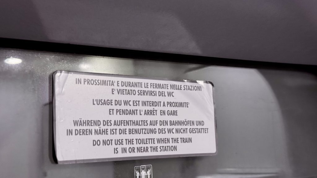 Ein Hinweisschild in mehreren Sprachen, das darauf hinweist, dass die Benutzung der Toilette im Zug in der Nähe von oder während des Haltens an Bahnhöfen verboten ist. Das Schild enthält Texte in Italienisch, Französisch, Deutsch und Englisch.