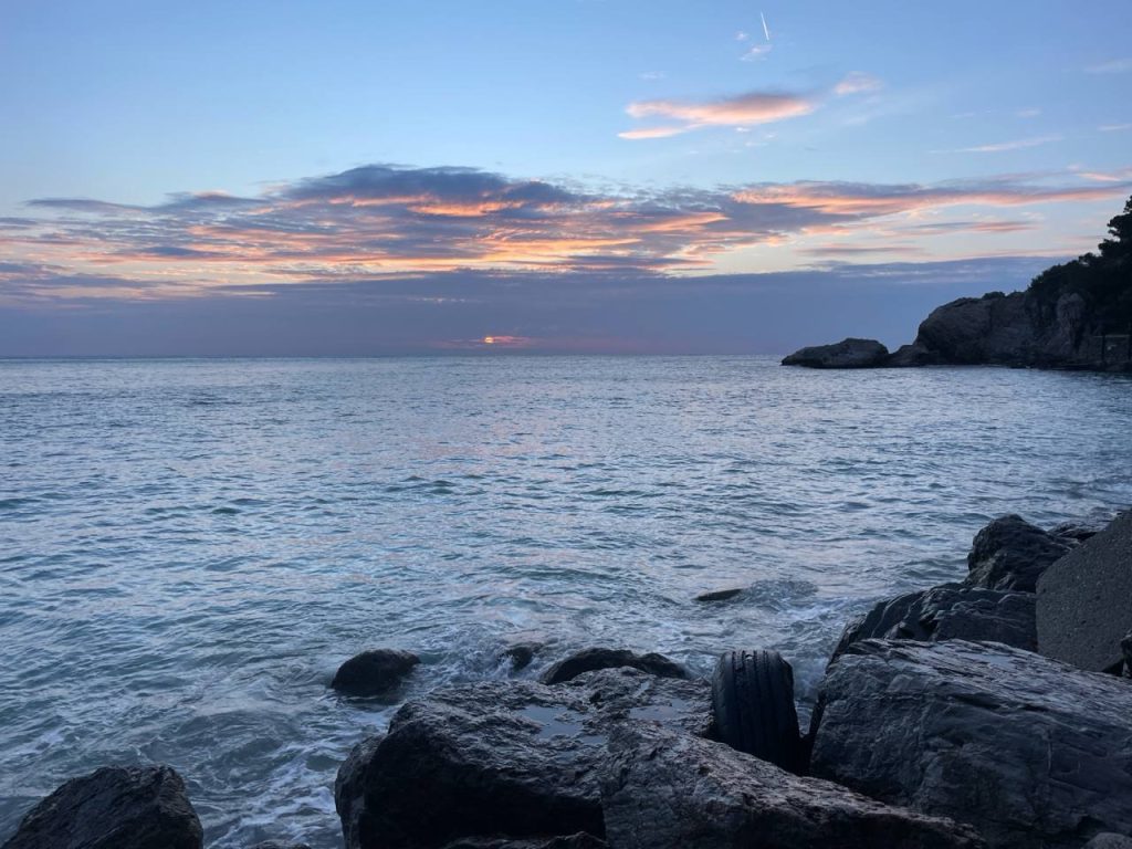Ein ruhiger Meereshorizont bei Sonnenuntergang, mit sanften Wellen, die gegen die Felsen im Vordergrund schlagen. Der Himmel ist von sanften Farbnuancen in Blau und Rosa durchzogen.
