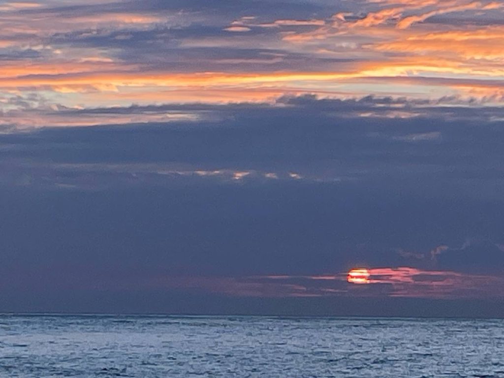Ein Sonnenuntergang über dem Meer, mit bunten Wolken in den Farben Orange, Pink und Lila, während die Sonne teilweise hinter der Wolkenfront verborgen bleibt. Die glatte Wasseroberfläche reflektiert die Farben des Himmels.